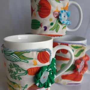 Trois tasses de thé habillées d'une housse de mug en tissu coton imprimé légumes (carottes, navets) et fleurs. Détail de finition avec bouton et fleur yo-yo artisanale verte.