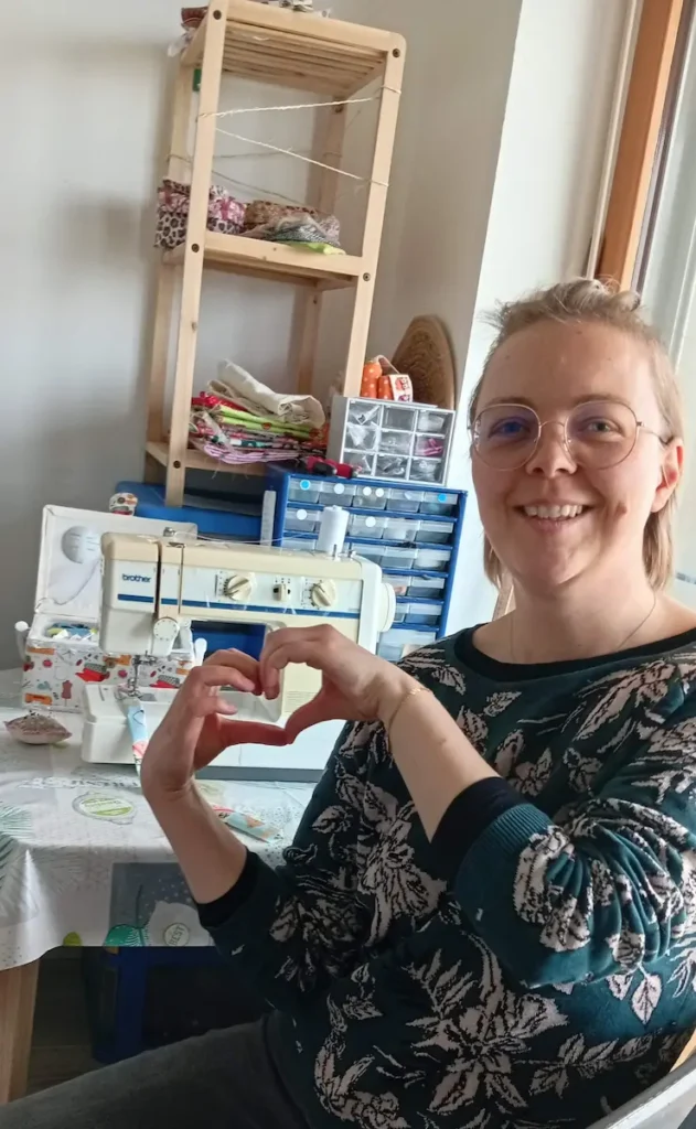 Créatrice de Cousu Mel qui fait un coeur avec les doigts devant sa machine à coudre et son espace de travail avec les tissus et tout le matériel de couture