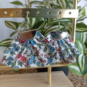 Boucles d'oreilles eventail motifs fleurs bleues et rouges sur fond blanc en tissu acier inoxydable Cousu Mel, accrochées sur un portant doré avec support en bois devant une plante verte