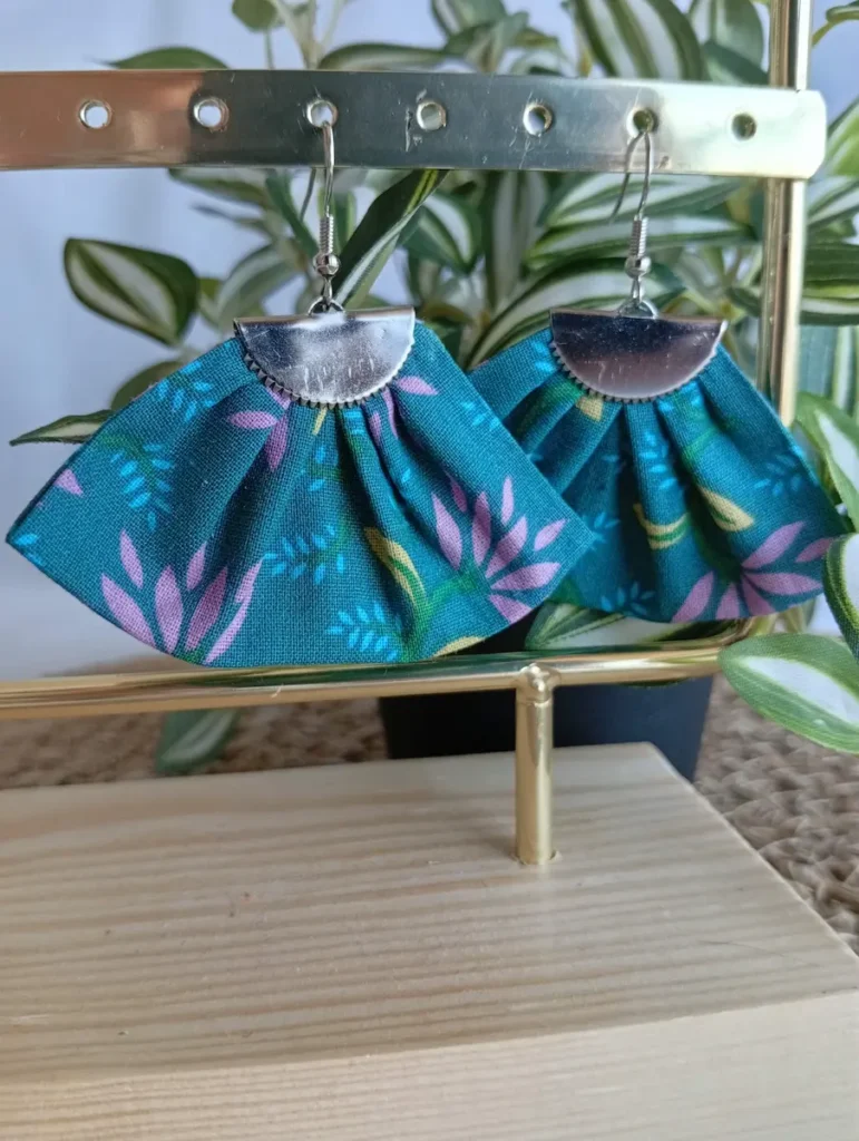 Boucles d'oreilles eventail motifs fleurs mauves et jaunes sur fond vert canard en tissu plissé et acier inoxydable Cousu Mel, accrochées sur un portant doré avec support en bois devant une plante verte