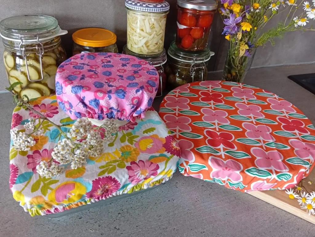 plan de travail anthracite sur lequel sont disposés trois plats recouverts d'une charlotte à fleurs aux motifs et couleurs assortis constituant un lot de charlottes couvre-plats "Boucle d'or". Au fond : des conserves maison de légumes colorés
