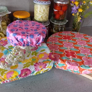 plan de travail anthracite sur lequel sont disposés trois plats recouverts d'une charlotte à fleurs aux motifs et couleurs assortis constituant un lot de charlottes couvre-plats "Boucle d'or". Au fond : des conserves maison de légumes colorés