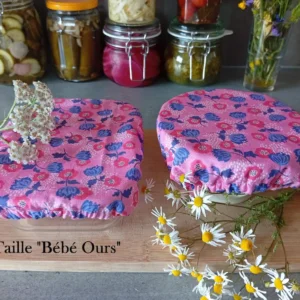 plan de travail avec 2 charlottes couvre-plats : l'une sur un bol, l'autre sur un petit plat rectangulaire. Au fond : des bocaux avec des légumes colorés et un bouquet de fleurs champêtre