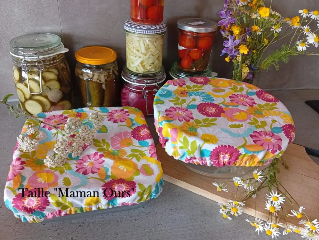 2 charlottes couvre-plats lavables sur un plan de travail : l'une recouvre un saladier moyen, l'autre un plat à gratin carré. Au fond : des bocaux de légumes colorés
