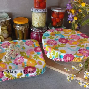 2 charlottes couvre-plats lavables sur un plan de travail : l'une recouvre un saladier moyen, l'autre un plat à gratin carré. Au fond : des bocaux de légumes colorés