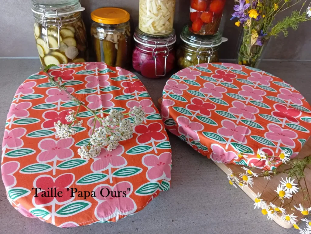 2 grandes charlottes couvre-plats motif à grosses fleurs roses sur fond orange disposées sur un plan de travail anthracite. L'un est sur un plat à tarte l'autre sur un grand plat à gratin. Au fond : des bocaux artisanaux de légumes au vinaigre