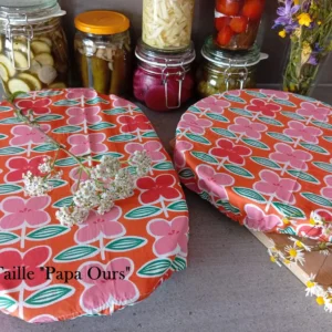 2 grandes charlottes couvre-plats motif à grosses fleurs roses sur fond orange disposées sur un plan de travail anthracite. L'un est sur un plat à tarte l'autre sur un grand plat à gratin. Au fond : des bocaux artisanaux de légumes au vinaigre