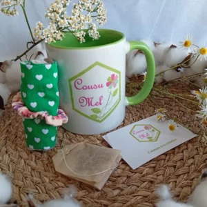 Coffret pause détente Cousu Mel comprenant un mug logo, une housse de tasse verte à motifs cœurs et un sachet d'infusion.