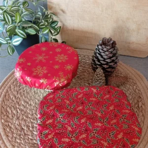 Deux plats couverts de 2 charlottes couvre-plats aux motifs de Noël, l'un flocons dorés sur fond rouge et l'autre houx doré sur fond rouge, reposent sur un set de table naturel ; à droite des pommes de pin, au fond une planche en bois, à gauche une plante verte