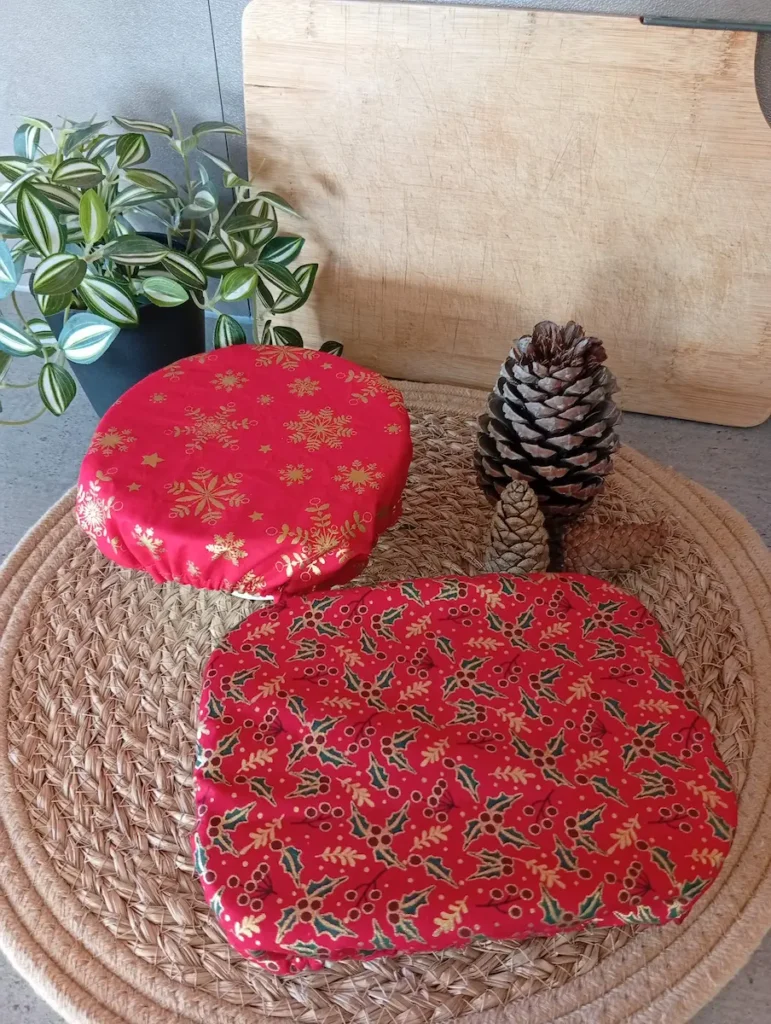 Deux plats couverts de 2 charlottes couvre-plats aux motifs de Noël, l'un flocons dorés sur fond rouge et l'autre houx doré sur fond rouge, reposent sur un set de table naturel ; à droite des pommes de pin, au fond une planche en bois, à gauche une plante verte