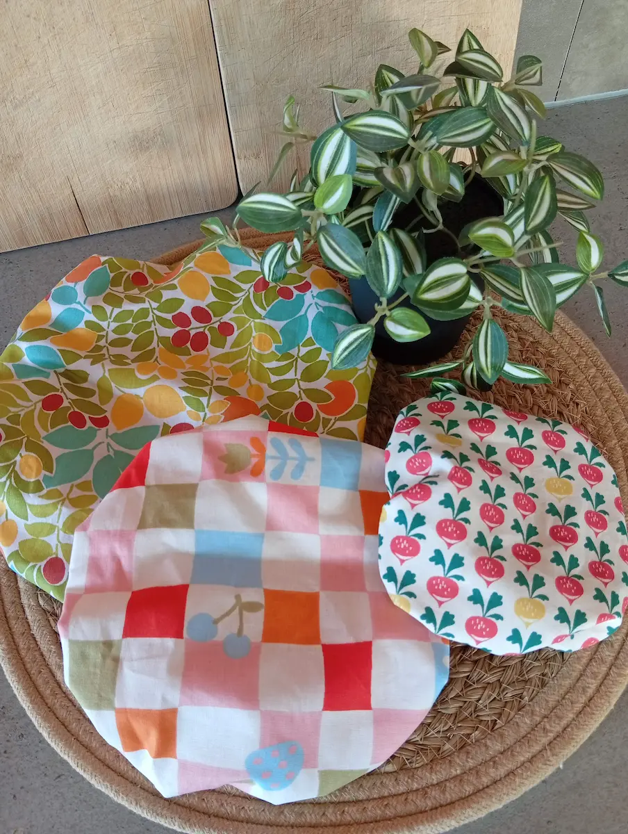lot de 3 tailles de charlottes couvre-plats avec trois motifs différents : citron, carreau cerise et betterave entourées d'une plante verte et de planche à découper en bois, posées sur un set de table rafia tressé