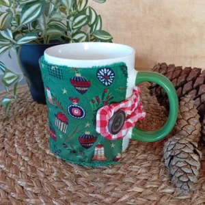 housse de tasse motifs de Noël : houx, cadeaux, cloche, décoration, boules, etc. sur fond vert ; autour du mug, il y a une plante verte et des pommes de pin sur un set de table naturel tressé