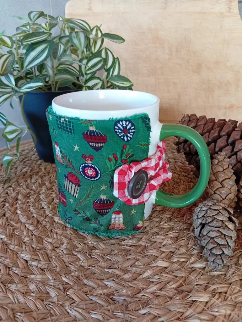 housse de tasse motifs de Noël : houx, cadeaux, cloche, décoration, boules, etc. sur fond vert ; autour du mug, il y a une plante verte et des pommes de pin sur un set de table naturel tressé